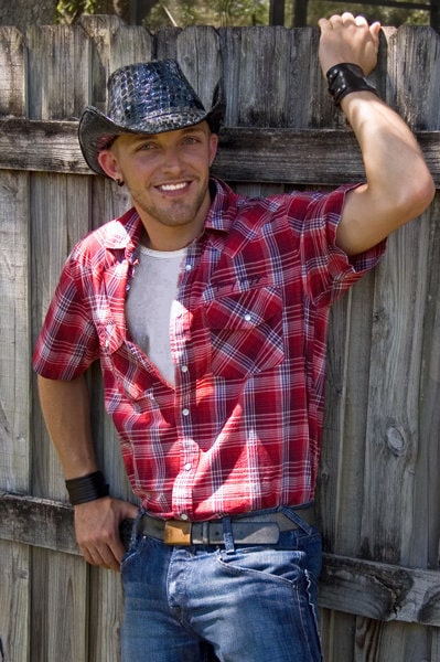 Christopher Pruitt, before his accident, standing in front of a fence smiling wearing a cowboy hat and red flannel shirt. 