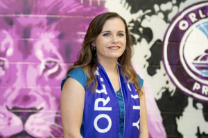 Danielle Head standing in front large painted mural, wearing blue drape around her neck that says "Orlando City"