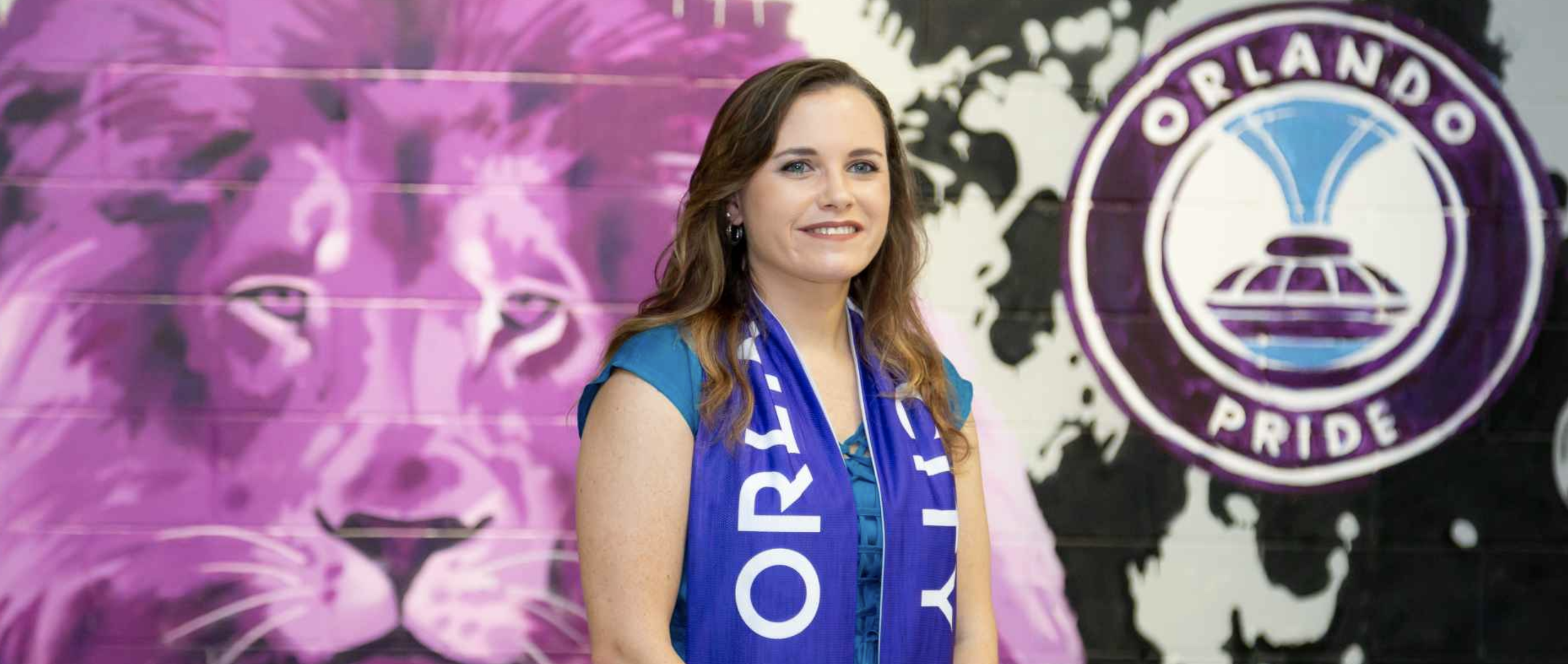 Danielle Head standing in front large painted mural, wearing blue drape around her neck that says "Orlando City"