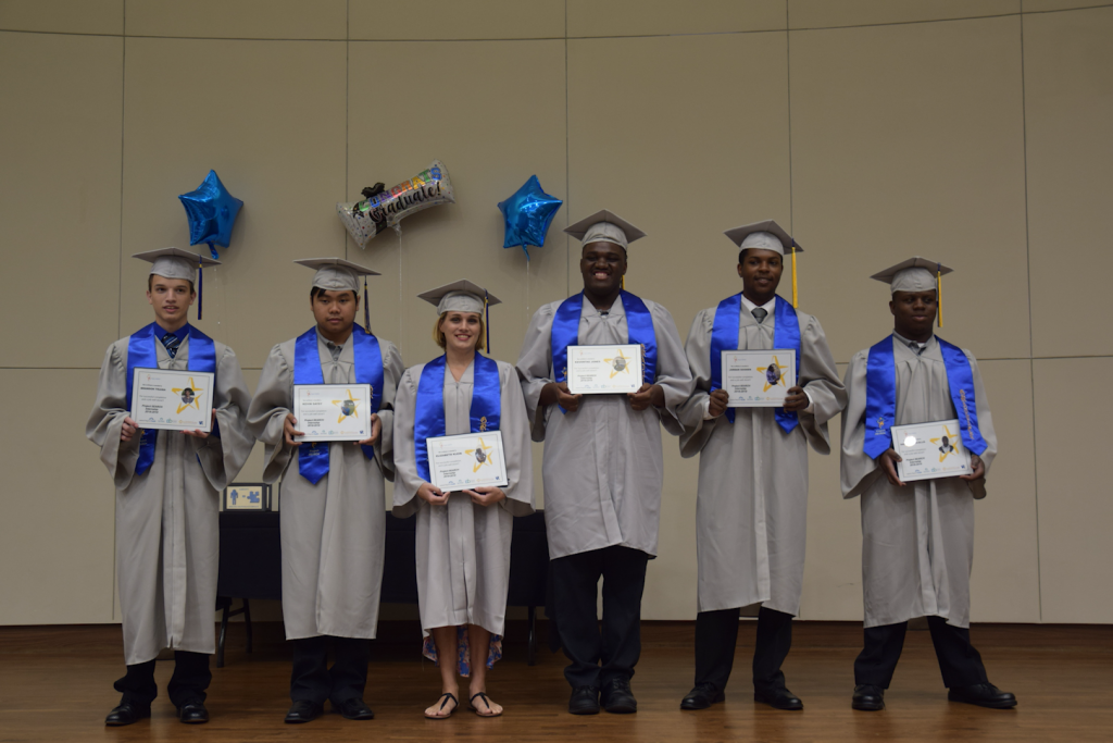 Six young individuals stand on stage wearing grey caps and gowns smiling and holding diplomas 