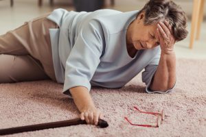 older woman laying on carpeted floor with hand on head nad look of pain on face. Pair of red reading glasses on floor, woman holding the top of walking cane.