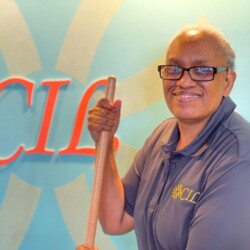 African American woman wearing square black glasses and smiling wears grey polo with yellow CIL logo. Woman holds the top of wooden broom and stands in front of indoor wall painted teal with orange CIL logo in center.