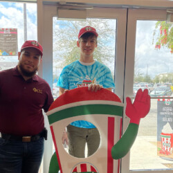 Standing inside of business in front of glass doors- two men. Man on left has beard and wears marron polo with yellow CIL logo. Younger man to right wears tie-dye blue shirt with Rita's ice logo and red Rita's hat, holding a Rita's sign.